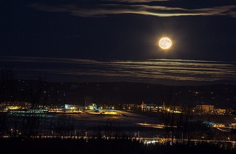 Image of a full moon hanging over UAF Campus, centered on West Ridge, and taken from far away. The image is predominantly dark blue, with orange lights in a line along the campus buildings. 