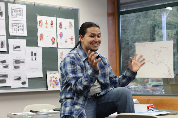 X'unei Lance Twitchell, a man wearing a plaid shirt, casually sitting on a desk teaching a Tlingit language class