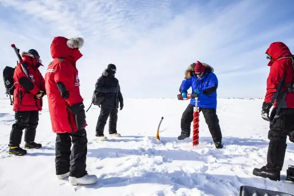 Four figures in heavy outdoor gear drill into ice amidst a snowy white expanse