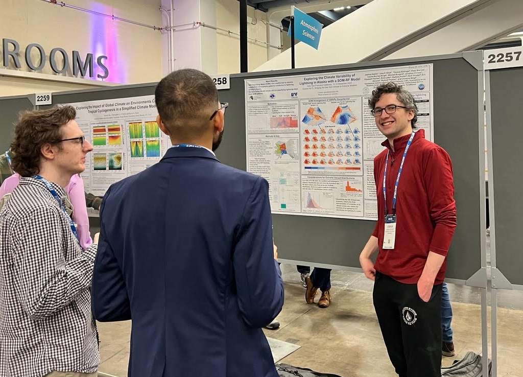 Two young men engage with Josh Hostler at his American Geophysical Union poster. Josh is smiling and wears a red shirt and plastic-rimmed glasses. 