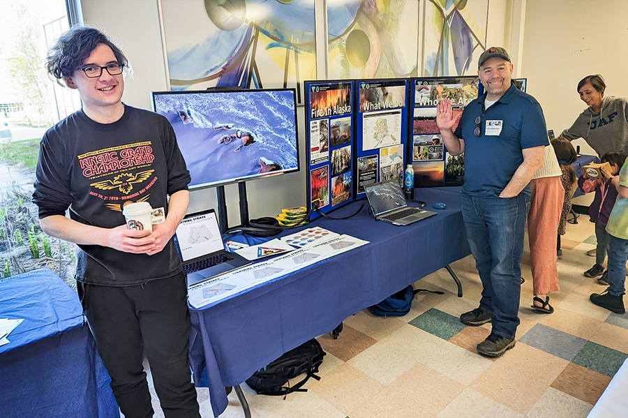 A tabling display with a computer monitor showing a map, and a poster board with information. Josh Hostler and Eric Stevens smile and wave at the camera.