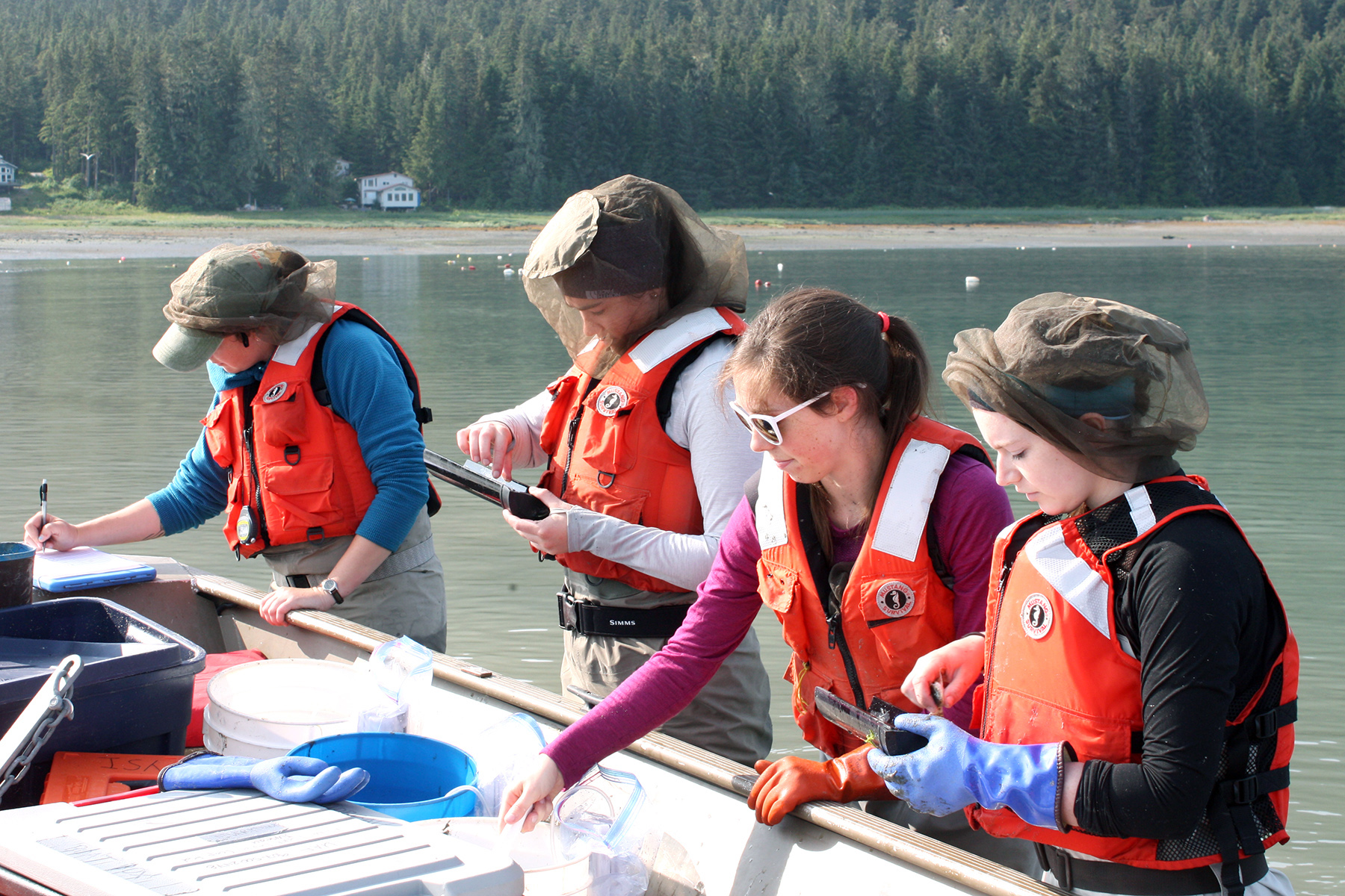 Female students in life jackets and mosquito head-nets record fish data on clipboards while standing beside a boat full of equipment.
