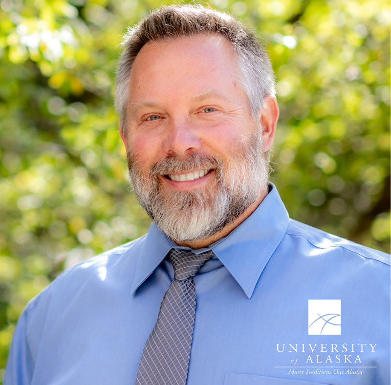 Brian Smentokski poses in front of a green tree, wearing a blue button up