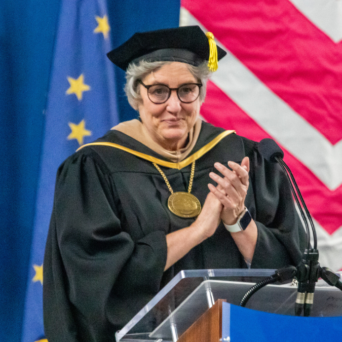 UA President Pat Pitney stands behind the podium and applauds with the audience at the 2023 UAF graduation